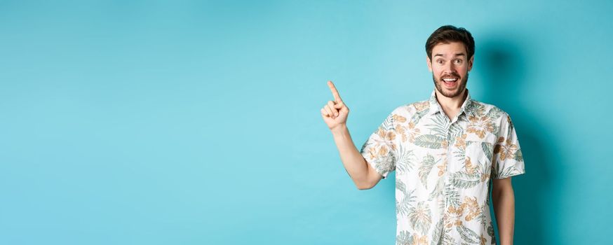 Surprised happy tourist pointing finger left at empty space, showing cool promo, standing in hawaiian shirt on blue background.