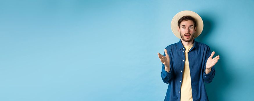 Shocked male tourist on vacation raising hands up and looking at camera startled, react to big news with disbelief, standing on blue background in straw summer hat and shirt.