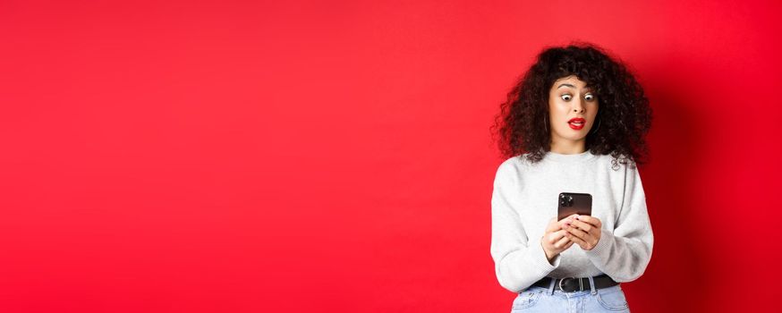 Shocked woman stare at smartphone screen with popped eyes, reading strange message, standing on red background.