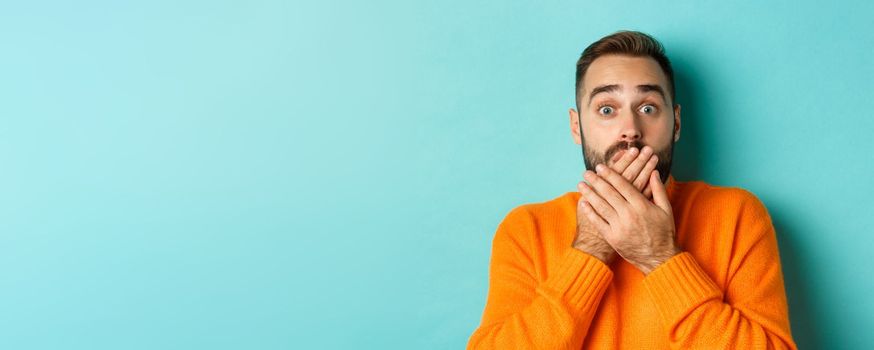 Close-up of shocked caucasian man covering his lips and mouth, staring astounded at camera, standing against turquoise background.