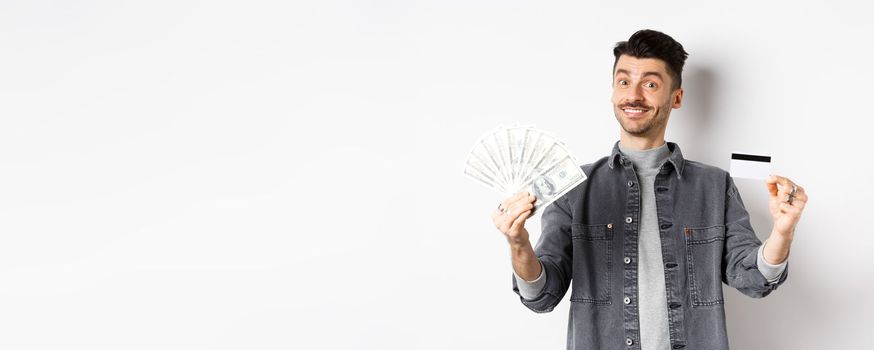 Happy guy showing plastic credit card and dollar bills, giving choice, standing on white background.
