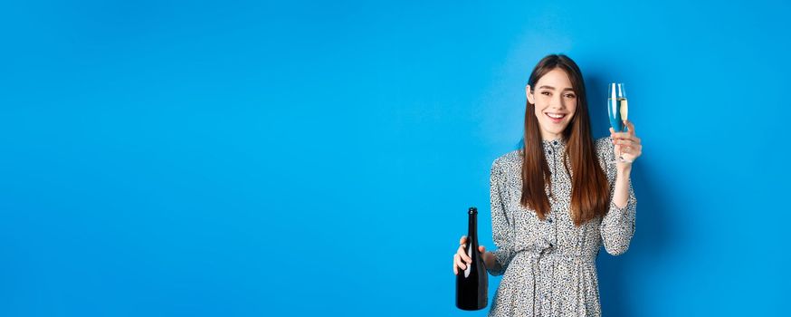 Celebration. Happy young woman in dress raising glass of champagne, holding bottle and smiling, having fun at party, standing on blue background.