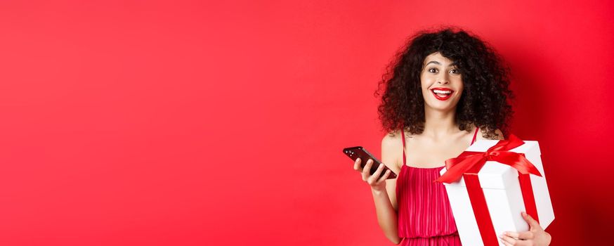 Online shopping and Valentines day. Beautiful young woman holding smartphone and lovers gift, looking surprised and happy at camera, red background.