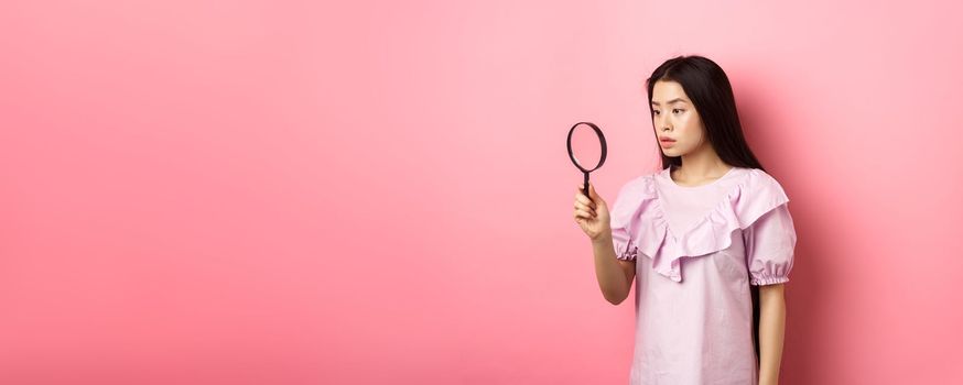 Serious girl investigating, looking through magnifying glass, searching for something, standing against pink background.