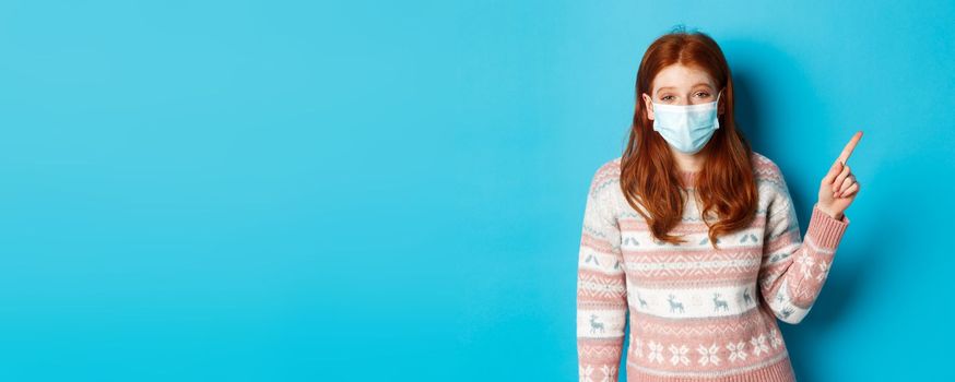 Winter, coronavirus and social distancing concept. Redhead girl in medical mask and sweater pointing at upper right corner and laughing, standing over blue background.