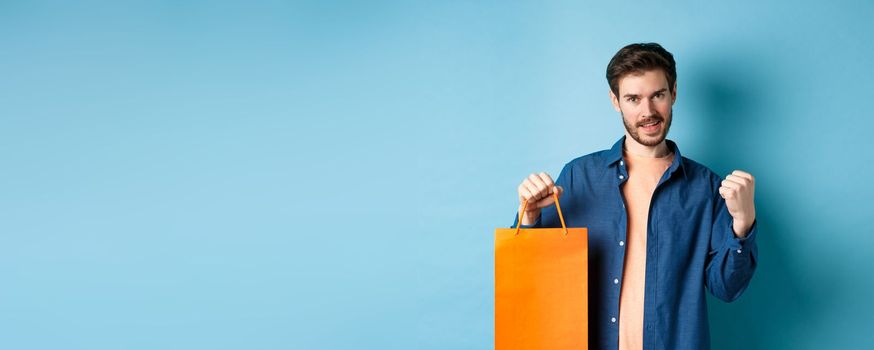 Handsome smiling man saying yes, triumphing and showing shopping bag, celebrate good promo offer, standing on blue background.