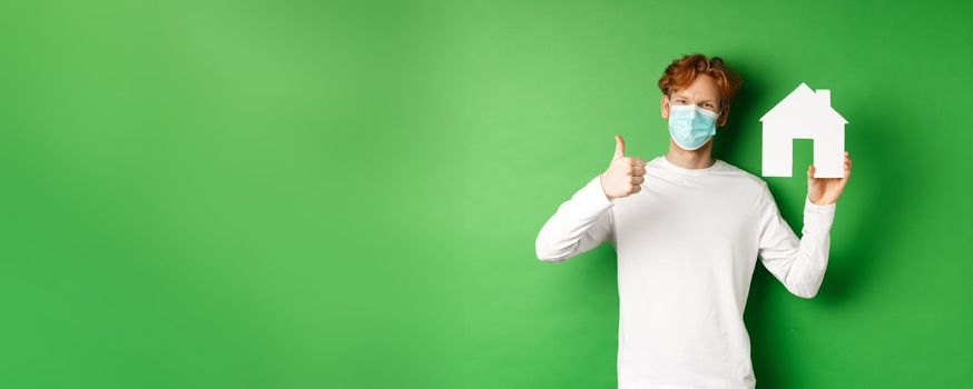 Real estate and covid-19 pandemic concept. Young redhead man in medical mask showing paper house cutout and showing thumb-up in approval, recommending agency.