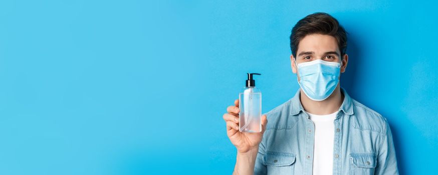 Concept of coronavirus, pandemic and preventive measures. Close-up of young man in medical mask advice to use hand sanitizer, showing antiseptic bottle, standing over blue background.