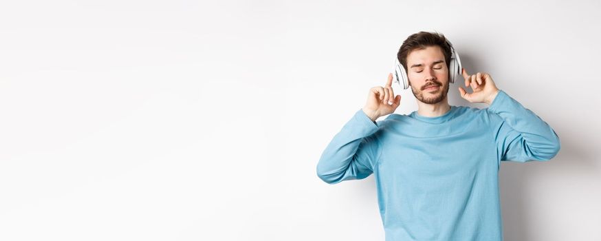 Relaxed young man enjoying favorite song, listening music in headphones with closed eyes and calm face, standing over white background.