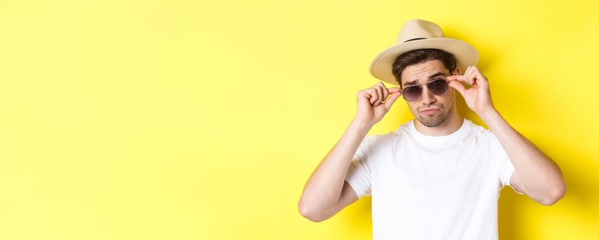 Concept of tourism and vacation. Close-up of cool tourist enjoying holidays on trip, wearing sunglasses with straw hat, yellow background.
