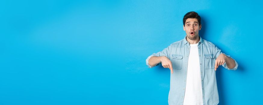 Surprised bearded adult man in casual outfit, pointing fingers down and looking startled, asking question about promotion, standing over blue background.