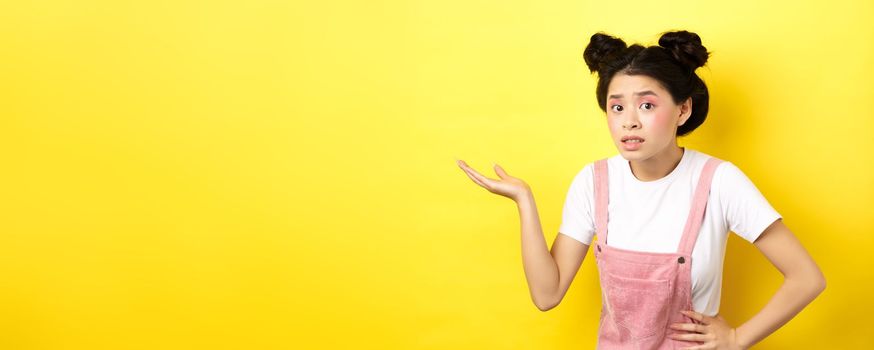 Confused asian teen girl with bright makeup, raising hand up clueless, looking puzzled, cant understand something, pointing at copy space, yellow background.