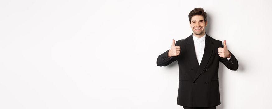 Concept of new year party, celebration and lifestyle. Image of attractive happy businessman in formal suit, showing thumbs-up and smiling, like and approve, standing over white background.