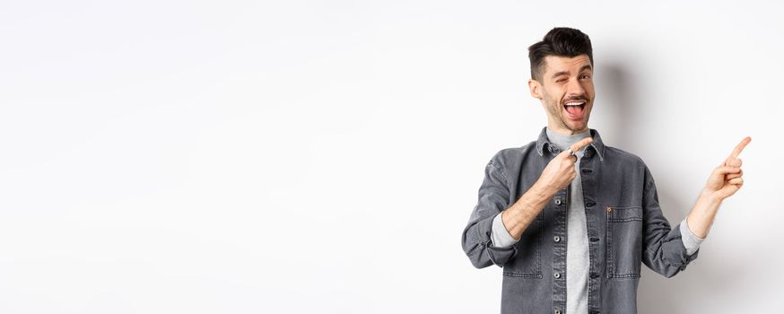 Handsome smiling guy making announcement, winking and pointing at upper right corner, showing logo, inviting to check out advertisement, standing on white background.