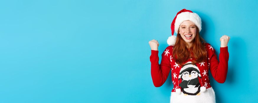 Happy holidays and Christmas concept. Cheerful redhead girl in santa hat and xmas sweater, winning and celebrating victory, raising hands up satisfied, triumphing.