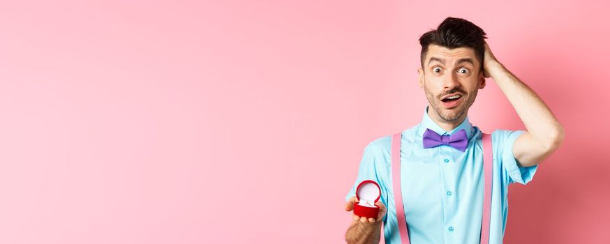 Valentines day. Confused funny guy standing with engagement ring, scratching head with unsure face, dont know what say, making proposal, standing over pink background.