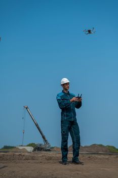 A man in a helmet and overalls controls a drone at a construction site. The builder carries out technical oversight