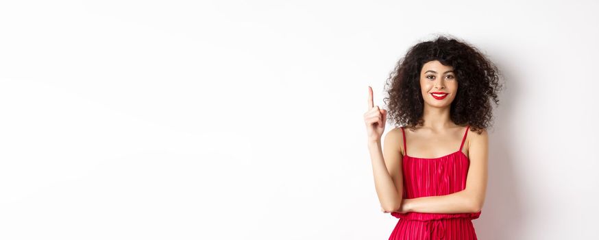 Beautiful smiling lady in red dress showing number one, raising finger and looking pleased, standing over white background.