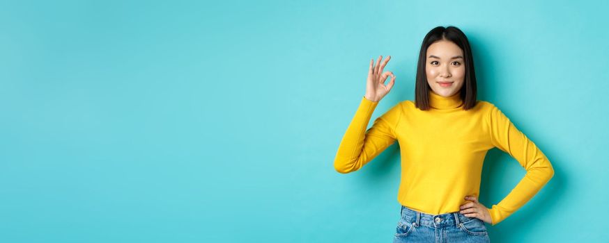 Beautiful smiling asian woman recommend product, showing Ok sign and looking satisfied, standing over blue background.