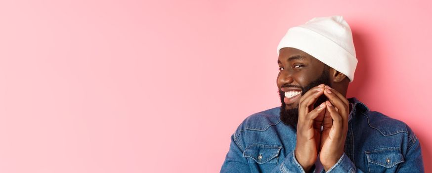 Close-up of devious african-american male model having an idea, scheming something, steeple fingers and smiling sly, standing over pink background.