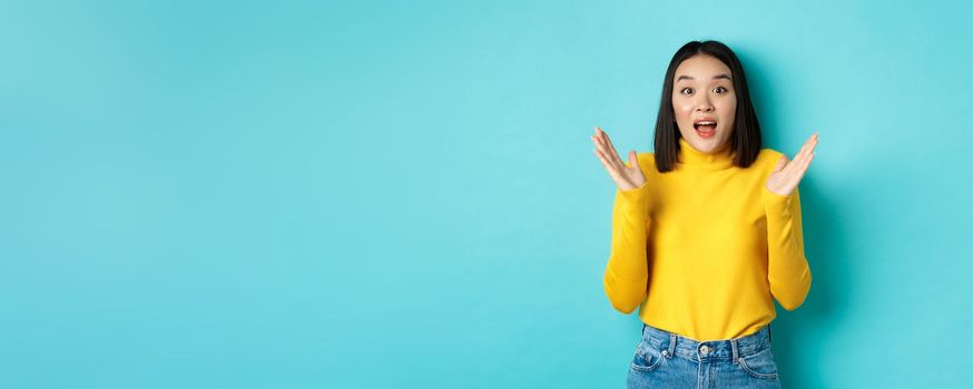 Image of happy asian woman clap hands and looking amazed, standing in yellow pullover over blue background.