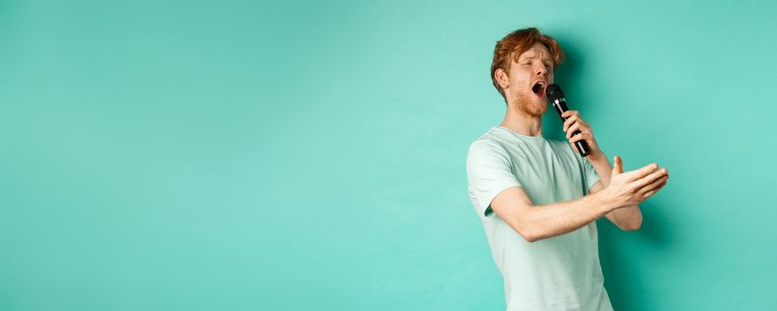 Passionate redhead man in t-shirt singing serenade with microphone, looking aside at karaoke and gesturing, standing over mint background.
