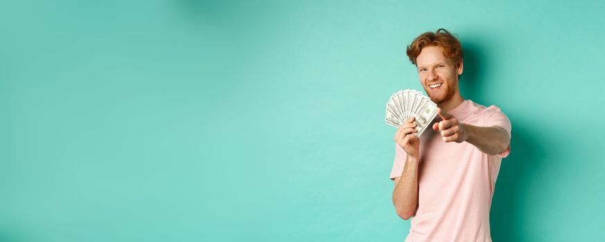 Handsome and confident redhead guy pointing finger at you, showing money dollars and smiling, concept of earning cash and investment.