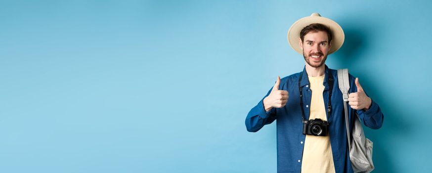 Cheerful handsome man recommending summer vacation place, showing thumbs up and smiling in approval. Tourist leave positive feedback, standing with camera and backpack on blue background.