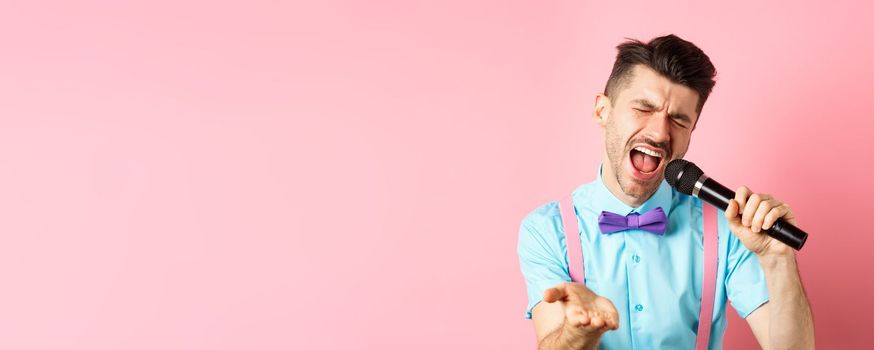 Party and festive events concept. Passionate singer holding microphone and pointing hand at you, singing about love, standing on pink background.