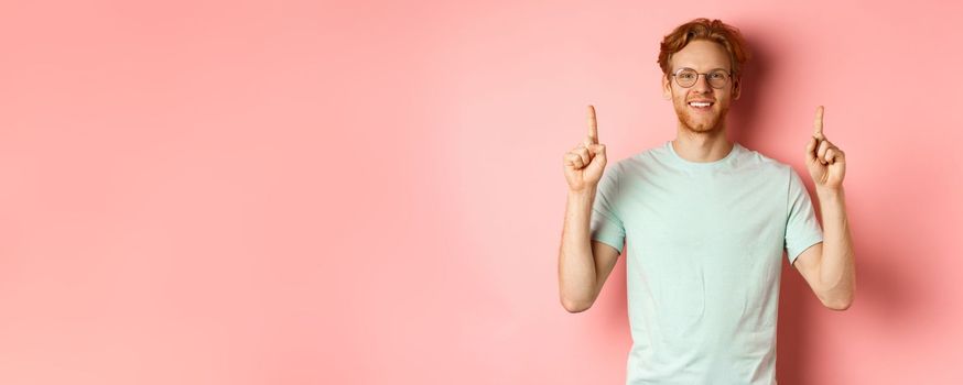 Attractive young man in glasses and t-shirt showing promo deal, pointing fingers up and smiling at camera, standing over pink background.
