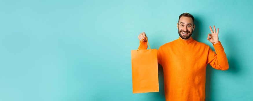 Satisfied male customer showing orange shopping bag and okay sign, recommending store, smiling pleased, standing over turquoise background.