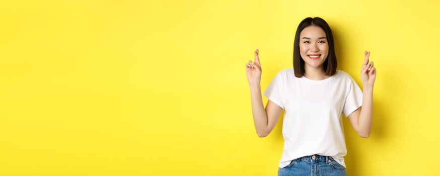 Hopeful asian girl making wish, cross fingers for good luck and praying with eyes closed, standing over yellow background.