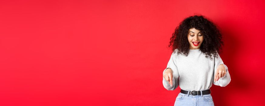 Dreamy beautiful woman with curly hair, pointing and looking down excited, checking out promo, standing against red background.