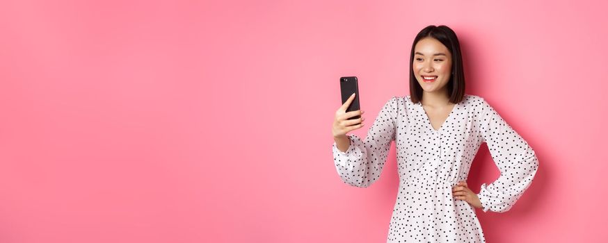 Beautiful asian girl using photo filters app and taking selfie on smartphone, posing in cute dress against pink background.