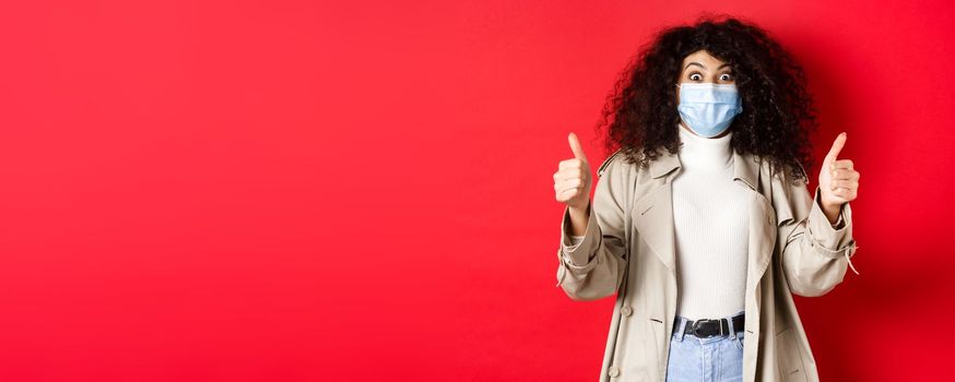 Covid-19, pandemic and quarantine concept. Excited girl with curly hair, wearing trench coat and medical mask, showing thumbs up in approval, red background.