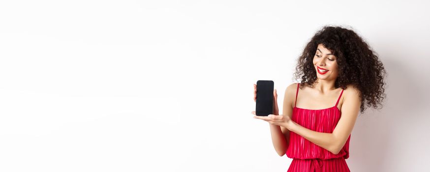 Beauty woman with makeup and curly hair, showing empty smartphone screen, demonstrate app, standing on white background.