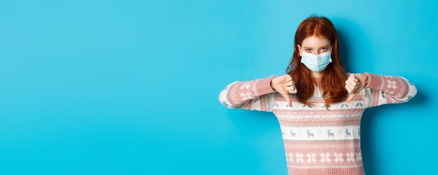 Winter, covid-19 and pandemic concept. Upset and angry redhead girl in face mask showing disapproval, thumbs down in dislike, standing over blue background.