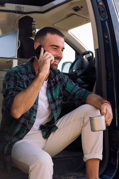 smiling young man talking on his mobile phone sitting in the door of his camper van drinking a mug of coffee, concept of freedom and digital nomad lifestyle