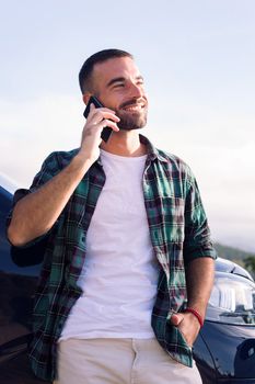 smiling young man talking on mobile phone leaning on his camper van, concept of freedom and nomad lifestyle