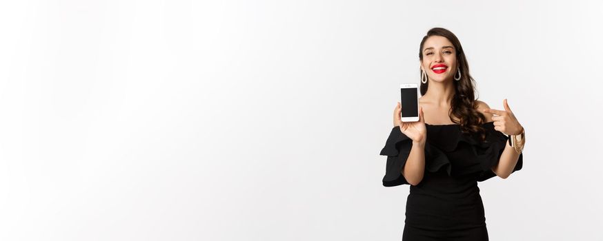 Online shopping concept. Fashionable woman in black dress pointing finger at smartphone screen, showing application, standing over white background.