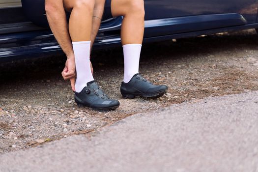 unrecognizable young man putting on cycling shoes sitting on his camper van to go training with his mountain bike, concept of freedom and sport in nature