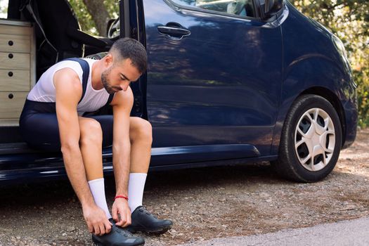 young man putting on cycling shoes sitting on his camper van to go training with his mountain bike, concept of freedom and sport in nature