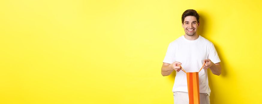Happy young man open shopping bag with present, smiling at camera, standing against yellow background.