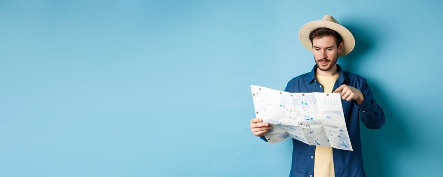 Handsome guy choosing place to go, pointing at sighseeing map on vacation, travelling on summer holidays, wearing straw hat and shirt, blue background.