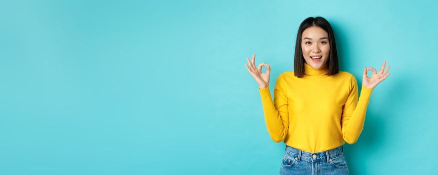 Cheerful asian female model showing okay gestures, smiling and looking impressed, praise product, standing over blue background.