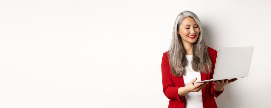 Business. Successful asian businesswoman in red blazer working on laptop, smiling and reading screen, standing over white background.