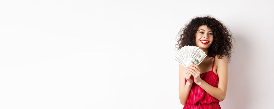 Excited woman in red dress winning money, showing dollar bills and smiling happy, standing on white background.