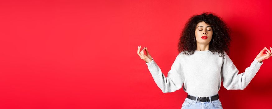 Woman in casual clothes trying to calm down with meditation, close eyes and practice yoga, holding hands sideways with zen gesture, standing on red background.