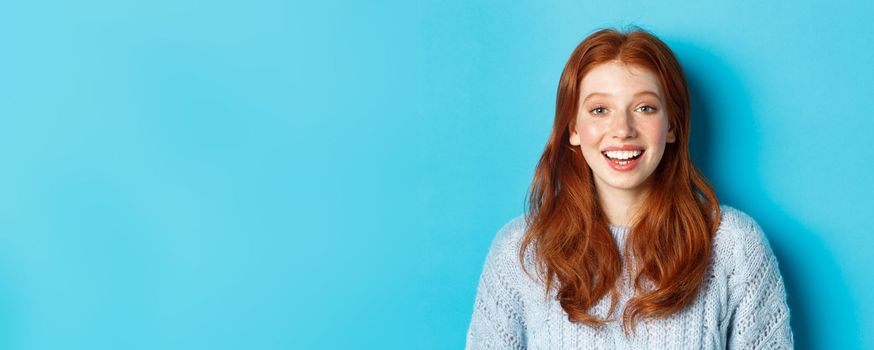 Close-up of happy redhead girl in sweater, looking at camera with hopeful smile, standing against blue background.