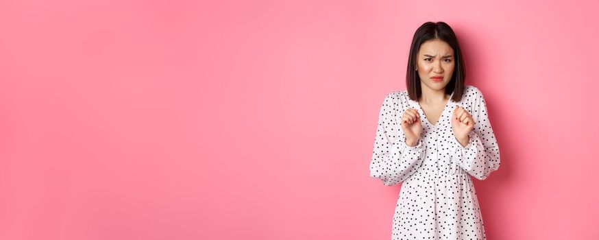 Disgusted asian woman staring with aversion and dislike, frowning and grimacing dissatisfied, standing in dress against pink background.